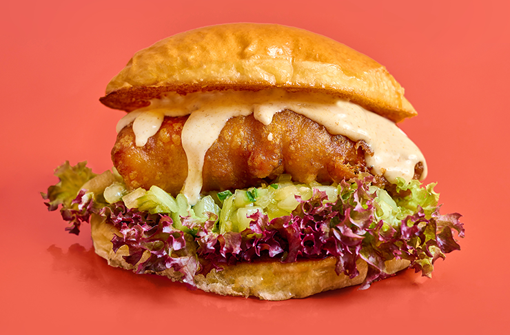 a deep fried moreton bay bug with mayo oozing down onto a bed lettuce in between a brioche bun with a red pastel background